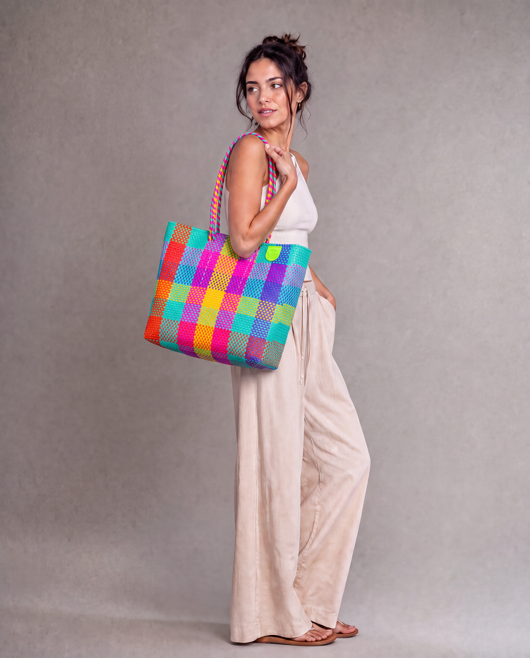 A woman wearing a Tin Marin woven tote bag with a colorful plaid pattern of pink, orange, yellow, green, blue, and purple against a neutral gray studio background. 