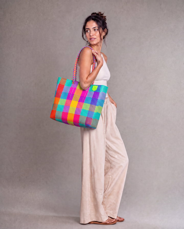 A woman wearing a Tin Marin woven tote bag with a colorful plaid pattern of pink, orange, yellow, green, blue, and purple against a neutral gray studio background. 