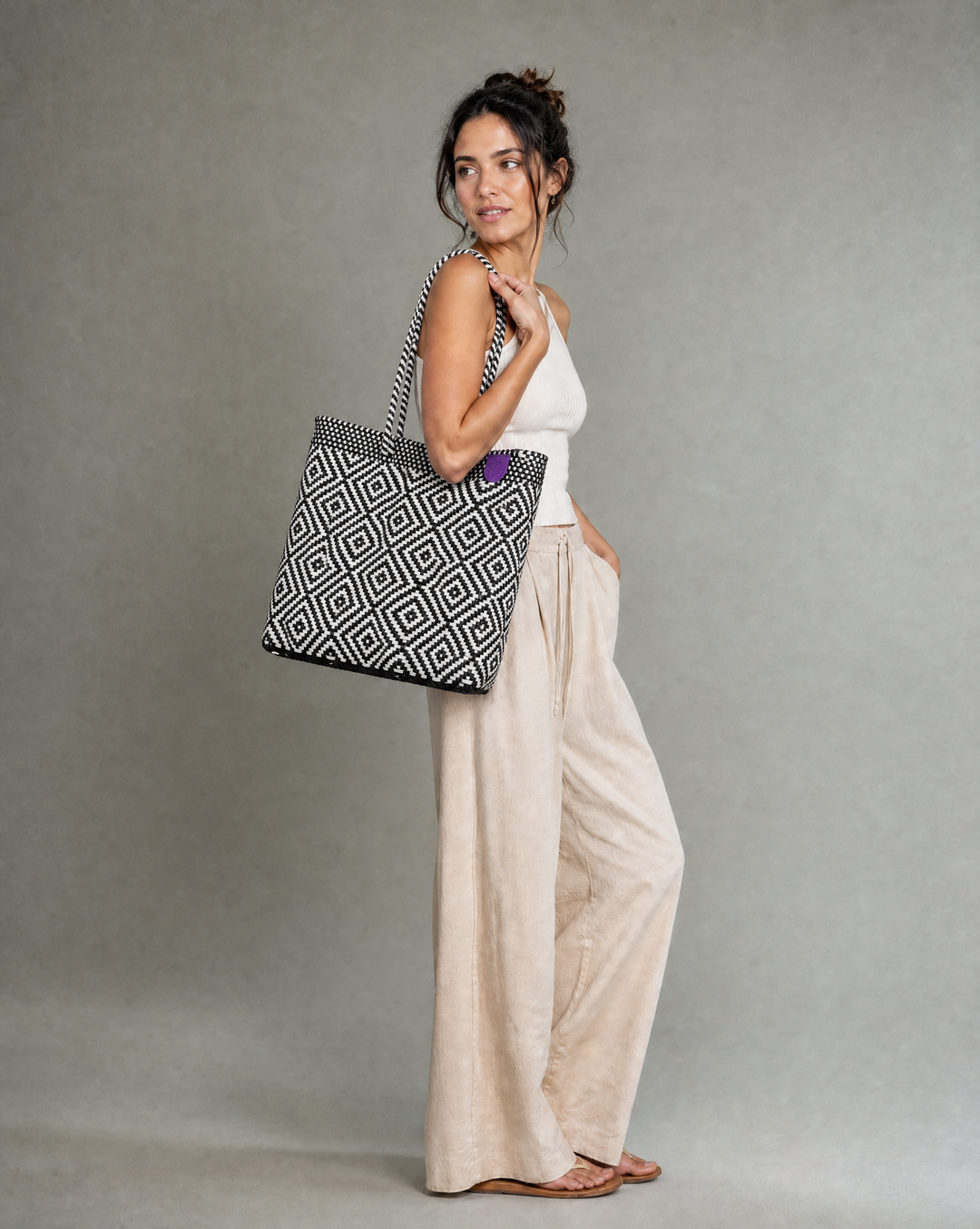 A woman wearing a Tin Marin woven tote bag with a rhombus pattern of black and white, against a neutral gray studio background. 