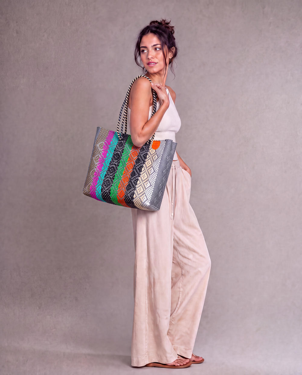 A woman wearing a Tin Marin woven tote bag with a striped pattern of multicolor against a neutral gray studio background. 