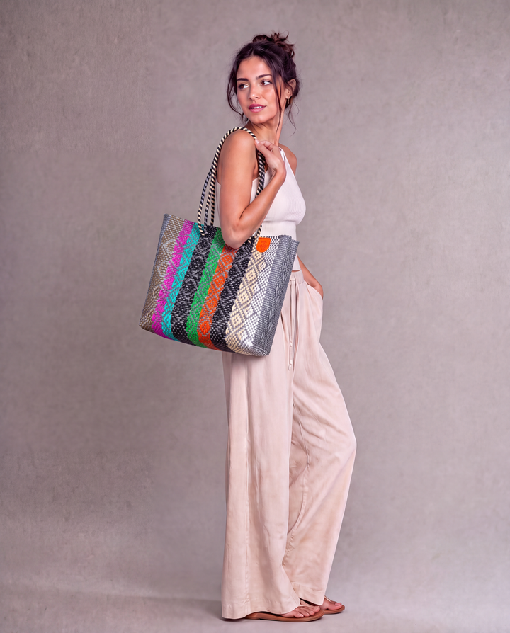 A woman wearing a Tin Marin woven tote bag with a striped pattern of multicolor against a neutral gray studio background. 