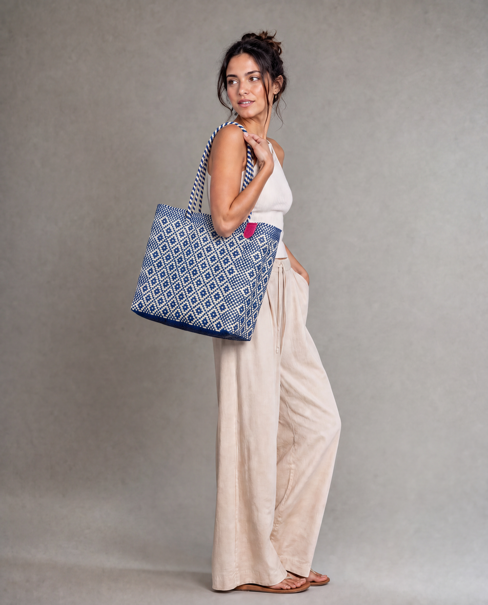 A woman wearing a Tin Marin woven tote bag with a rhombus pattern of blue and beige, against a neutral gray studio background. 